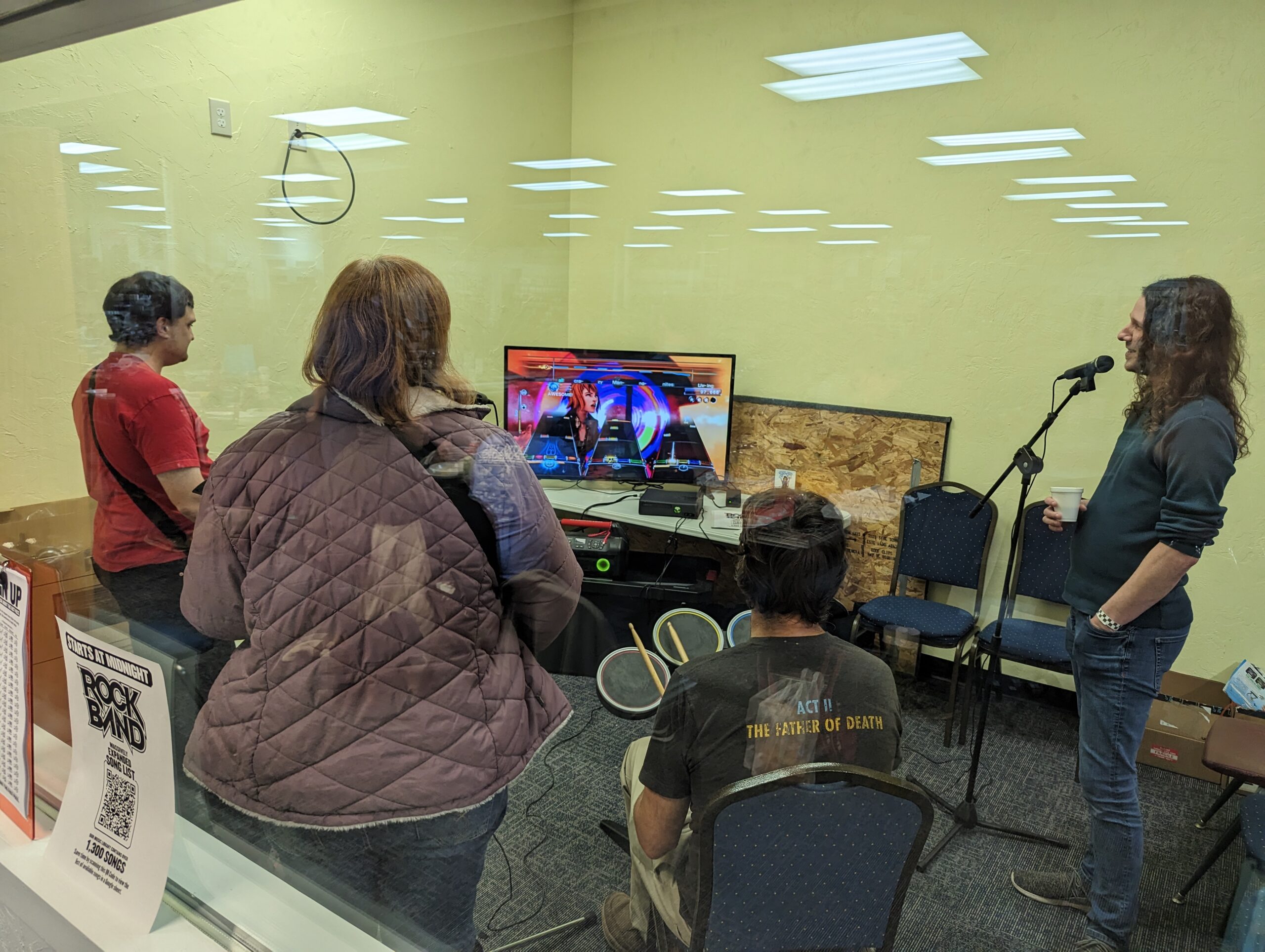 non-profit-09 Four people play the game Rock Band with various instruments and a song on TV.